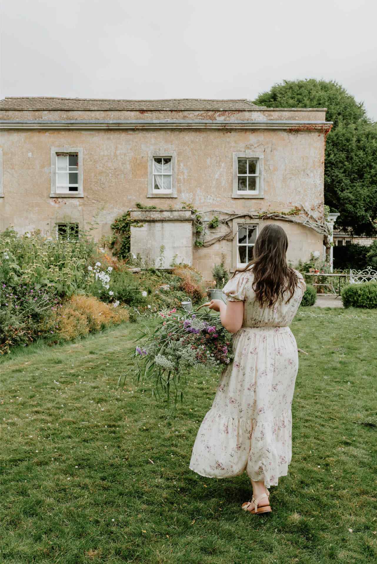 A woman in a floral summer dress holds a trug of flowers in front of a Cotswolds stone house