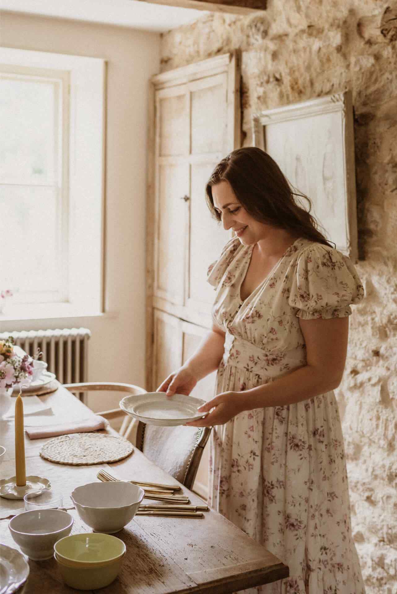 A woman in a floral dress and long dark hair lays a table in an elegant Georgian dining room